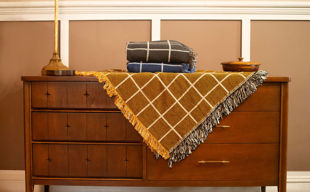 Greer Acorn golden brown grid woven throw displayed on a wooden dresser with blue and charcoal Greer blankets