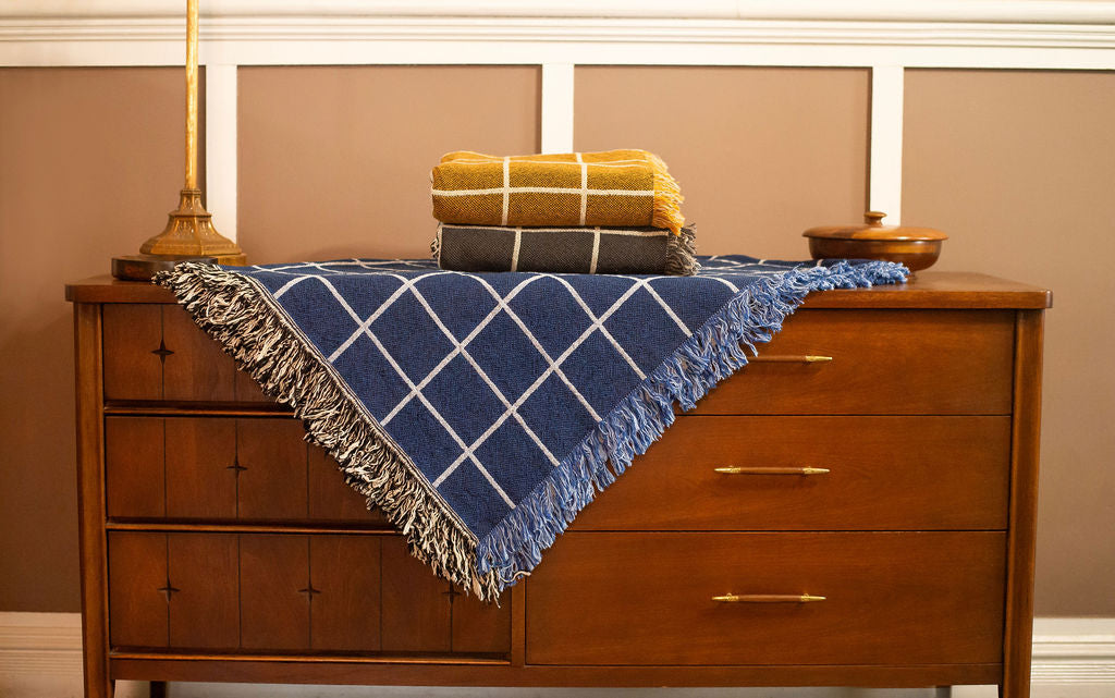 Greer Lapis blue grid woven throw displayed on a wooden dresser with gold and charcoal Greer blankets