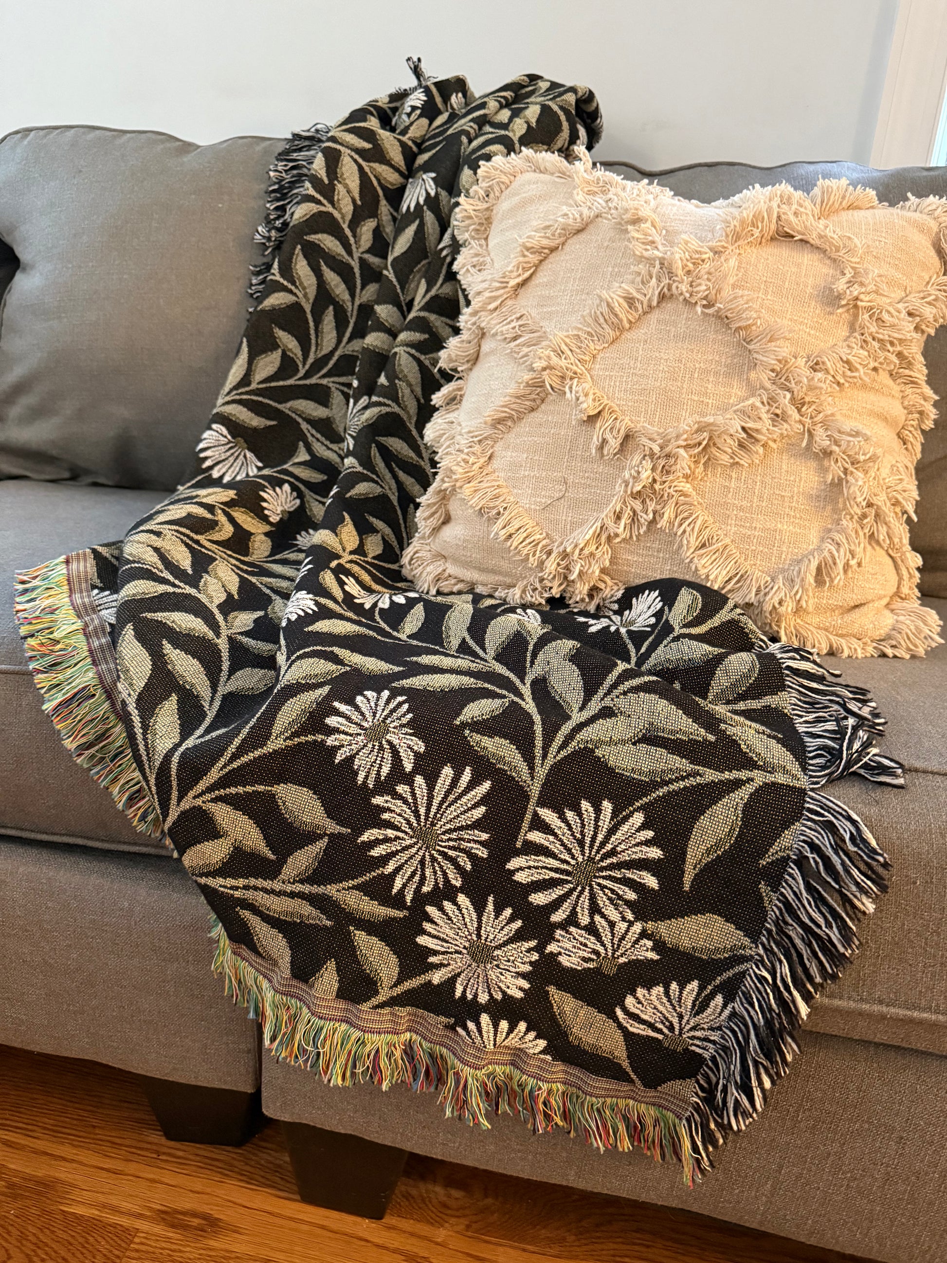 May Bramble botanical throw blanket draped over a gray sofa beside a textured cream pillow